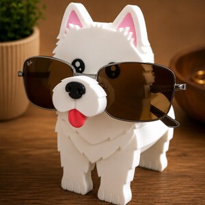 May include: A white dog-shaped sunglasses holder with pink-lined ears and a red tongue. The holder is wearing brown sunglasses. The dog is standing on a wooden surface. A small plant is in the background.