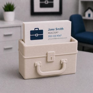 May include: A beige business card holder in the shape of a briefcase, containing a stack of white business cards. The cards display a blue briefcase icon and the name "Jane Smith, REALTOR&reg;", with contact details.