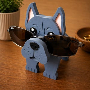 May include: A blue wooden dog-shaped eyeglass holder with a black nose and pink inner ears. The holder is wearing a pair of brown-tinted sunglasses. The holder is on a wooden surface.
