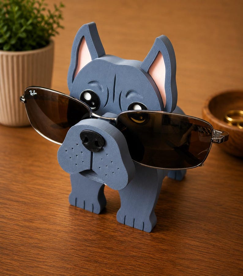 May include: A blue wooden dog-shaped eyeglass holder with a black nose and pink inner ears. The holder is wearing a pair of brown-tinted sunglasses. The holder is on a wooden surface.