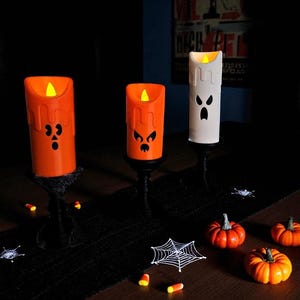 May include: Halloween-themed decorative candles in orange and white, featuring carved faces and flickering LED lights. The candles are set on black stands, with miniature pumpkins, spiderwebs, and candy corn scattered around.