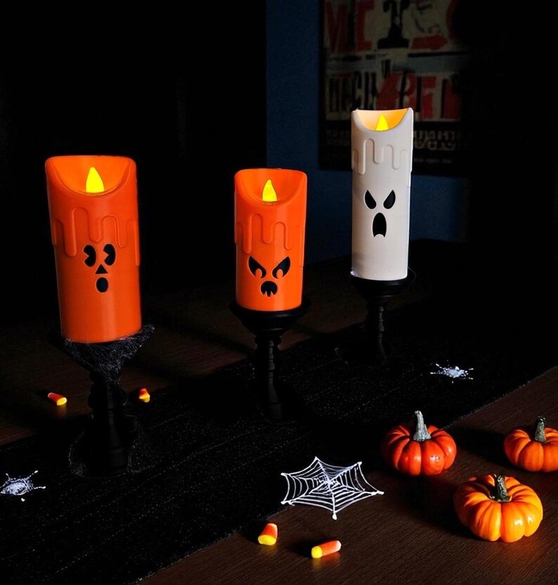 May include: Halloween-themed decorative candles in orange and white, featuring carved faces and flickering LED lights. The candles are set on black stands, with miniature pumpkins, spiderwebs, and candy corn scattered around.