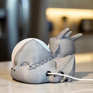 Puede incluir: Un soporte de carga para tel&eacute;fono con forma de dinosaurio gris. El soporte tiene una superficie texturizada y un cable blanco que sale de la base. El dinosaurio tiene una cabeza redondeada y una espalda puntiaguda, con un puerto de carga en el centro.