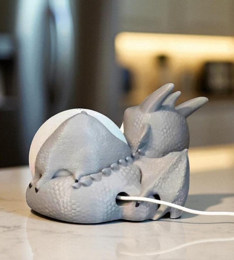 Puede incluir: Un soporte de carga para tel&eacute;fono con forma de dinosaurio gris. El soporte tiene una superficie texturizada y un cable blanco que sale de la base. El dinosaurio tiene una cabeza redondeada y una espalda puntiaguda, con un puerto de carga en el centro.