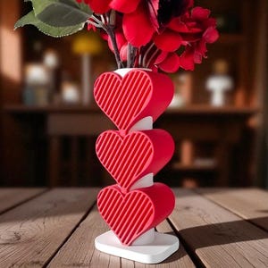 K&ouml;nnte beinhalten: Eine dekorative Vase mit einem einzigartigen Design mit drei gestapelten roten Herzformen mit Streifenmuster. Die Vase enth&auml;lt rote Blumen und steht auf einem wei&szlig;en Sockel. Die Vase steht auf einem Holztisch.