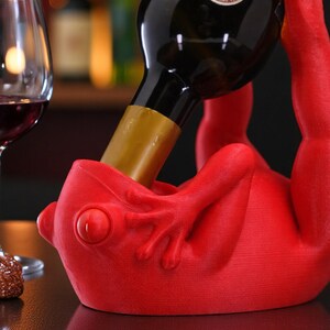May include: A red frog-shaped wine bottle holder supporting a wine bottle. The holder has a detailed frog design. A wine glass with red wine is in the background. The holder is made of a solid material.