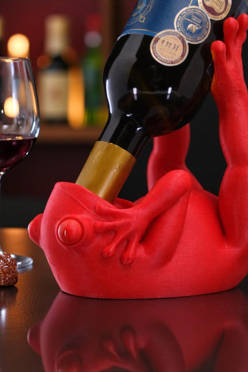 May include: A red frog-shaped wine bottle holder supporting a wine bottle. The holder has a detailed frog design. A wine glass with red wine is in the background. The holder is made of a solid material.