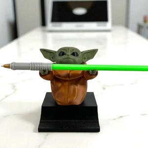 May include: A 3D-printed Grogu figure, in a brown robe, holding a green lightsaber-shaped pen. The figure is on a black rectangular stand. The pen is grey with a gold tip. The background is a blurred office setting.