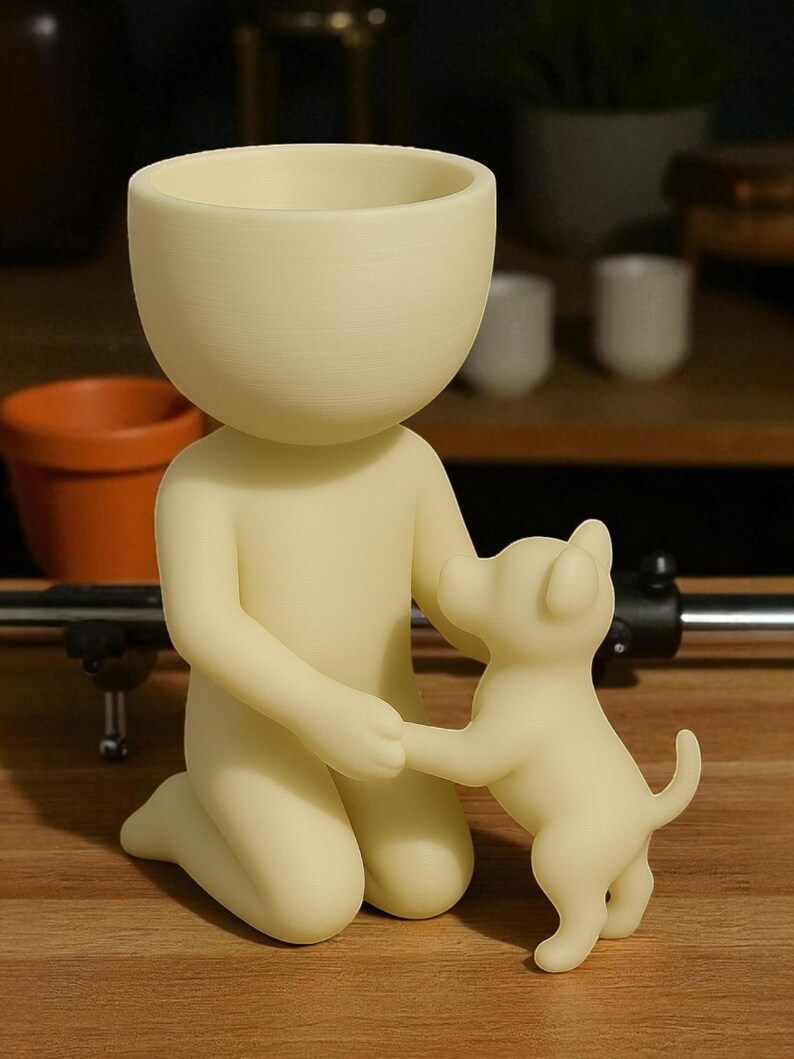 K&ouml;nnte beinhalten: Ein cremefarbener Pflanztopf in Form einer knienden Person, die die Pfote eines kleinen Hundes h&auml;lt. Die Person hat einen gro&szlig;en, schalenf&ouml;rmigen Kopf, perfekt f&uuml;r Pflanzen. Der Hund steht ebenfalls cremefarben auf den Hinterbeinen. Der Pflanztopf steht auf einer Holzoberfl&auml;che.