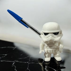 May include: A white Stormtrooper figurine holding a blue and clear pen. The figurine has black eye coverings and is standing on a black and white surface. The pen is angled towards the upper left.