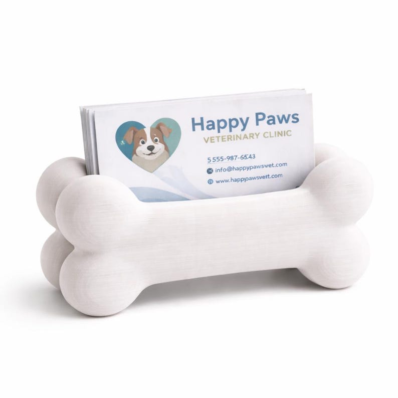K&ouml;nnte beinhalten: Ein wei&szlig;er, knochenf&ouml;rmiger Visitenkartenhalter. Der Halter enth&auml;lt einen Stapel Visitenkarten mit dem Text "Happy Paws VETERINARY CLINIC" und einem Logo eines Hundes in einem Herzen. Kontaktinformationen sind ebenfalls sichtbar.