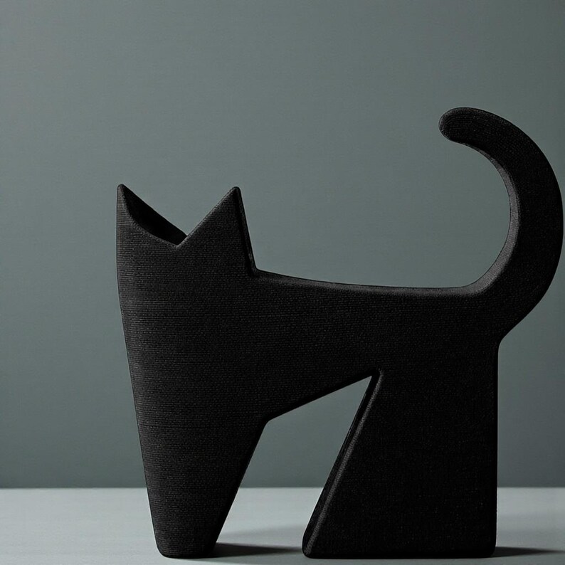 May include: A black cat-shaped decorative object with a textured surface. The cat has a pointed ear and a curved tail. The object is made of a solid material and stands on a light-colored surface.