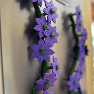 May include: Purple flower magnets with green centers and stems are attached to a metallic surface. The flowers are in full bloom and arranged in a vertical pattern. The background is blurred, highlighting the floral design.