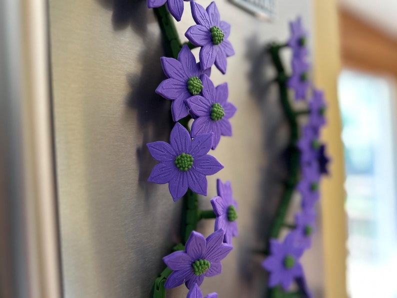 May include: Purple flower magnets with green centers and stems are attached to a metallic surface. The flowers are in full bloom and arranged in a vertical pattern. The background is blurred, highlighting the floral design.