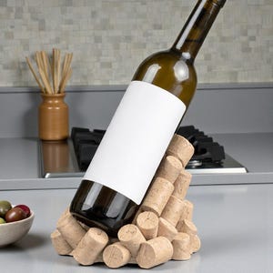 May include: A green wine bottle with a blank white label is displayed in a cork wine holder. The holder is made of stacked corks. A small bowl of olives and a vase with sticks are in the background.