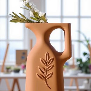 May include: Terracotta-colored ceramic vase with a handle and a leaf design. The vase holds white flowers and green leaves. The vase is a decorative item for the home.