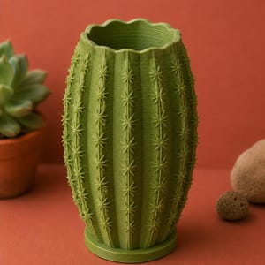 May include: A green cactus-shaped vase with a textured surface and star-shaped details. The vase has a scalloped rim and sits on a round base. The background is a solid reddish-orange color.