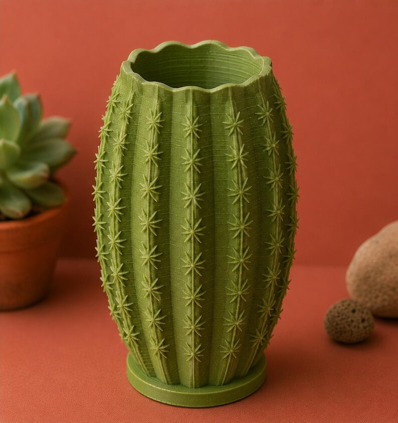 May include: A green cactus-shaped vase with a textured surface and star-shaped details. The vase has a scalloped rim and sits on a round base. The background is a solid reddish-orange color.