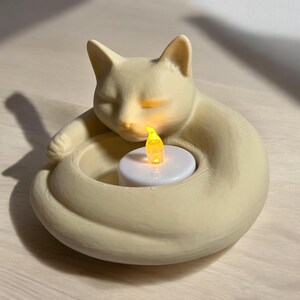 May include: A cream-colored cat-shaped candle holder with a sleeping cat design. The cat's body forms a circular shape, holding a white candle with a flickering yellow LED light. The candle holder is made of a smooth material.