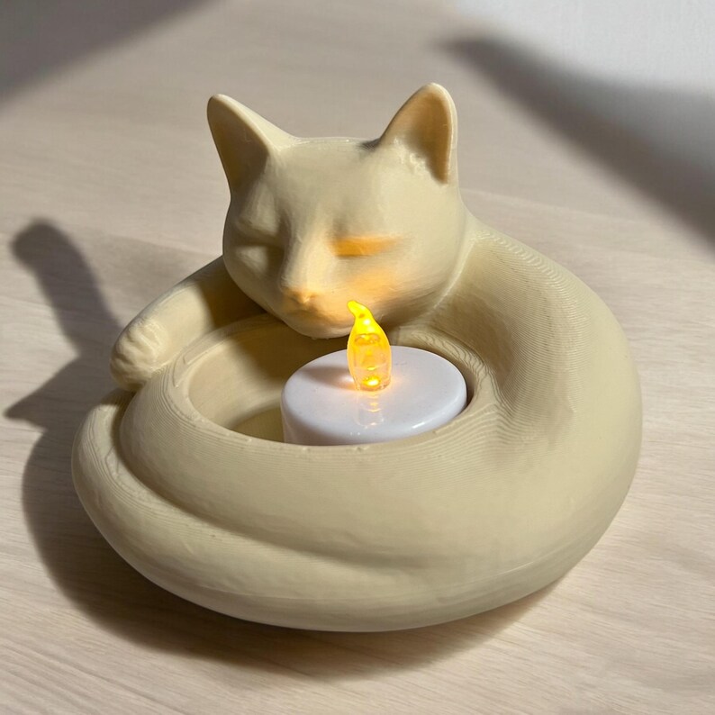 May include: A cream-colored cat-shaped candle holder with a sleeping cat design. The cat's body forms a circular shape, holding a white candle with a flickering yellow LED light. The candle holder is made of a smooth material.