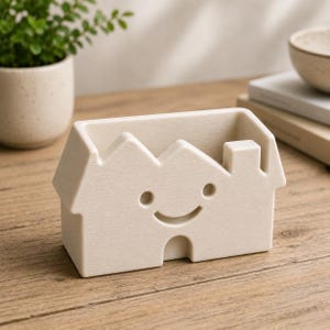 May include: A beige ceramic house-shaped desk organizer with a smiling face. The organizer has a rectangular opening for storage and a chimney detail. The house is sitting on a wooden surface.