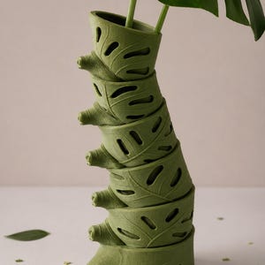 Puede incluir: Un jarr&oacute;n verde apilado con un dise&ntilde;o en forma de hoja, que sostiene una planta de monstera. El jarr&oacute;n est&aacute; compuesto por varias secciones en forma de hoja, cada una con detalles recortados. Las grandes hojas verdes de la planta contrastan con la forma del jarr&oacute;n.