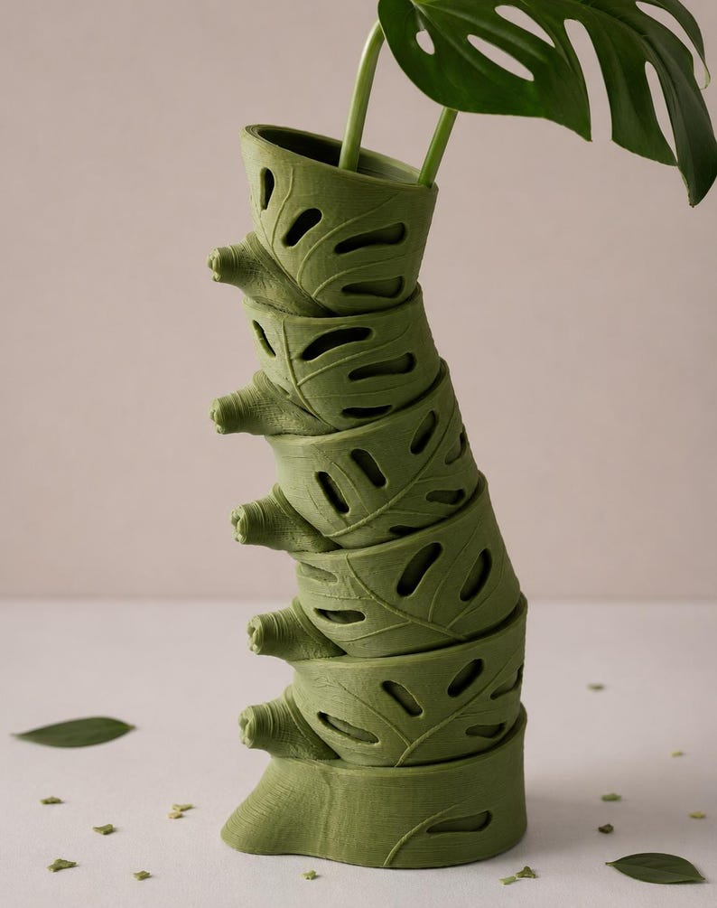 Puede incluir: Un jarr&oacute;n verde apilado con un dise&ntilde;o en forma de hoja, que sostiene una planta de monstera. El jarr&oacute;n est&aacute; compuesto por varias secciones en forma de hoja, cada una con detalles recortados. Las grandes hojas verdes de la planta contrastan con la forma del jarr&oacute;n.