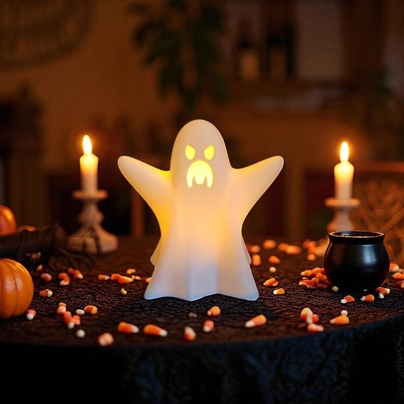 May include: A glowing white ghost-shaped Halloween decoration with outstretched arms and a carved face. The ghost is illuminated from within, sitting on a black tablecloth with candy corn scattered around. Lit candles and a small cauldron are also present.