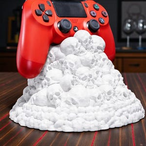 Può includere: Un controller di gioco rosso poggia su un supporto bianco a forma di teschio. Il controller ha pulsanti neri e pad direzionali. Il supporto è un cumulo di piccole e dettagliate teschi. Lo sfondo include un tavolo di legno e una libreria.