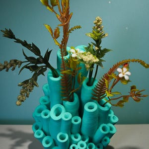 May include: A turquoise, coral-shaped vase with various openings, holding an assortment of artificial plants and flowers. The plants feature green, brown, and white accents, creating a vibrant contrast against the vase's color.
