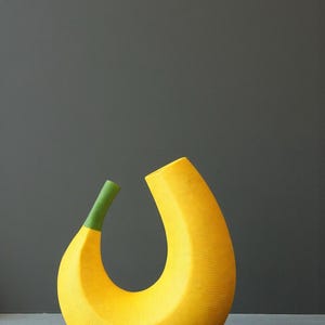 May include: A large, yellow, banana-shaped ceramic vase with a green stem. The vase has a textured surface and a curved, open design. The background is a solid gray.
