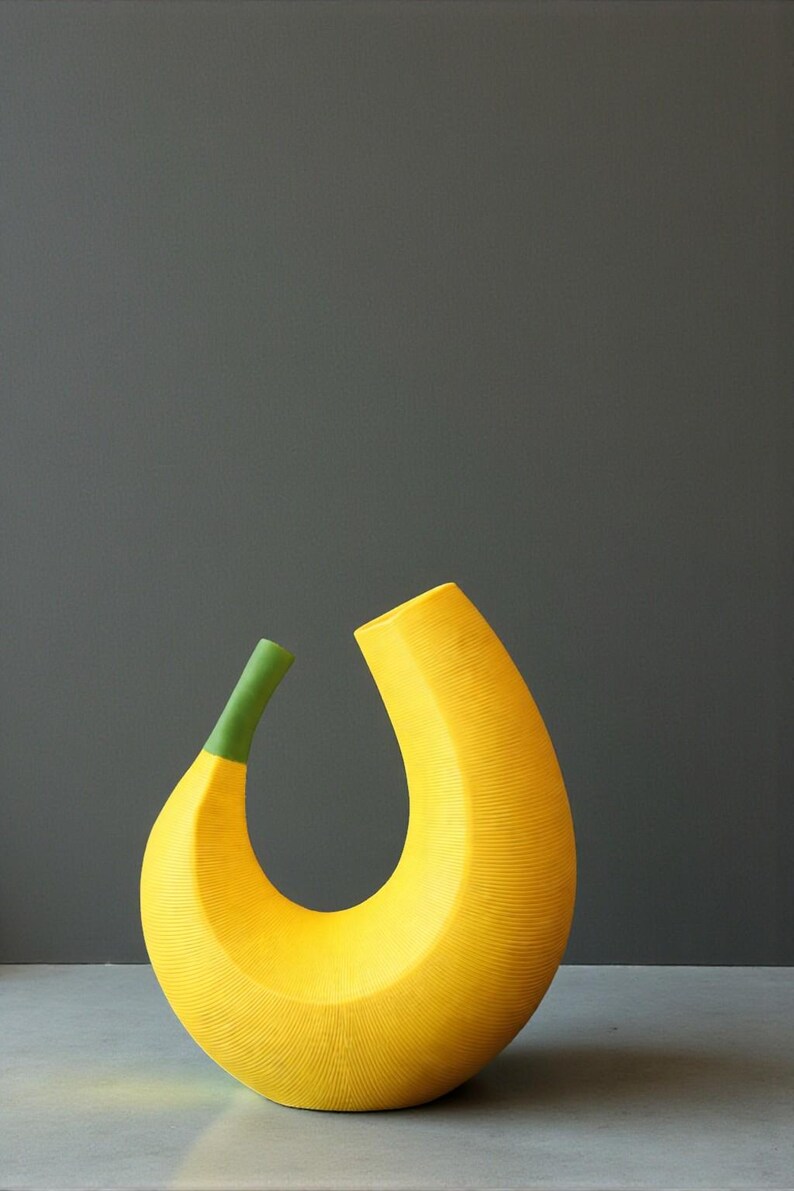 May include: A large, yellow, banana-shaped ceramic vase with a green stem. The vase has a textured surface and a curved, open design. The background is a solid gray.