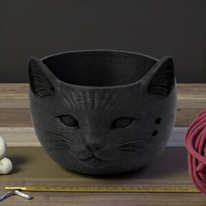 May include: A black cat-shaped planter with a textured surface. The planter features a detailed cat face with ears, eyes, nose, and whiskers. Two small holes are on the side of the face. The planter is designed to hold small plants or other decorative items.