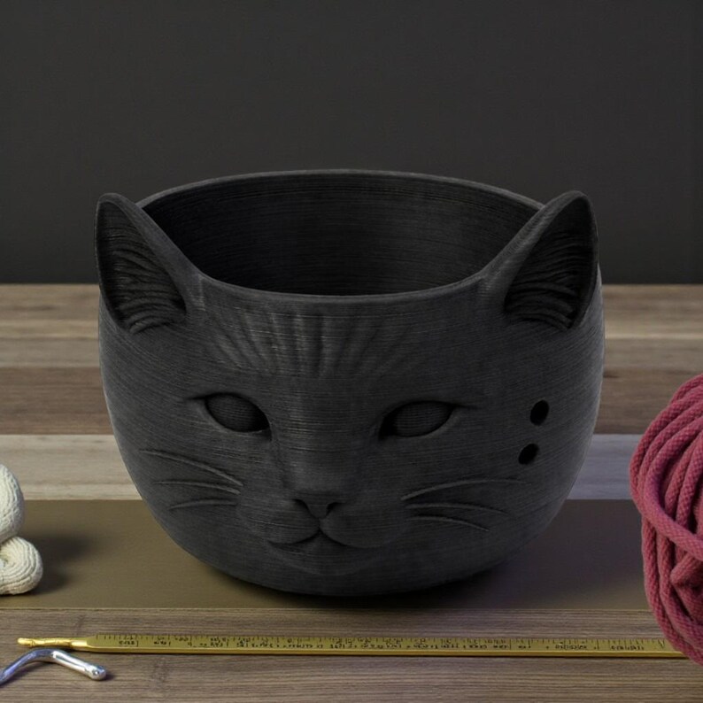 May include: A black cat-shaped planter with a textured surface. The planter features a detailed cat face with ears, eyes, nose, and whiskers. Two small holes are on the side of the face. The planter is designed to hold small plants or other decorative items.