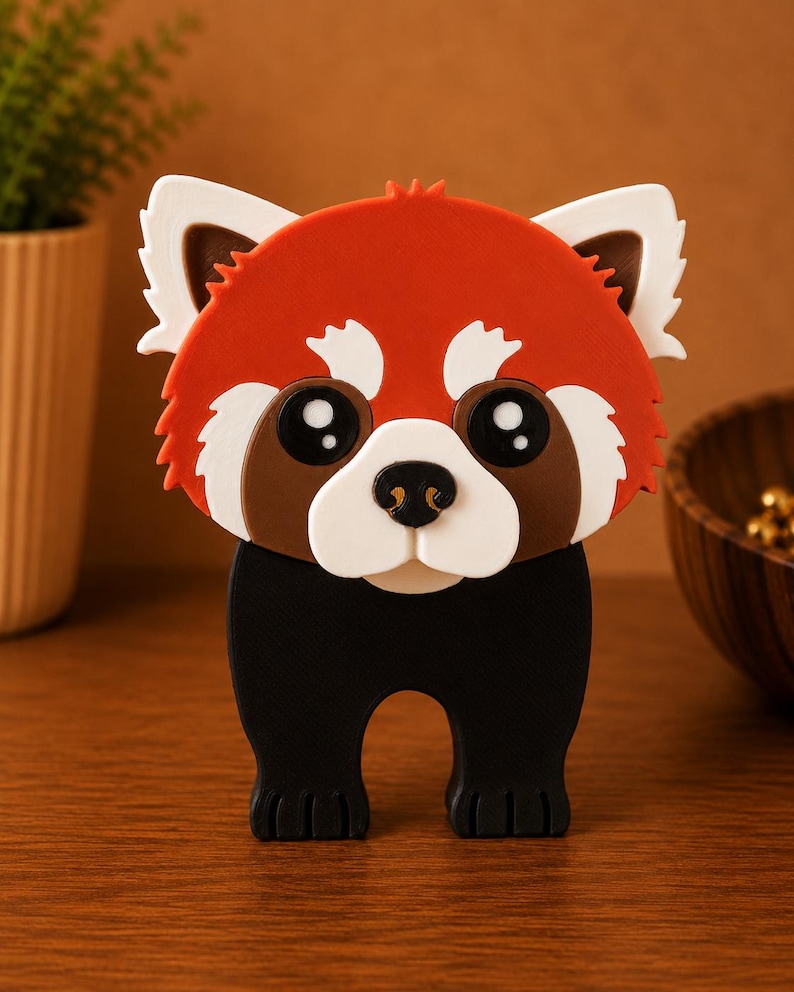 May include: A whimsical red panda figurine with a detailed design. The figurine features a red head with white and brown accents, large black eyes, and a black body. It is standing on a wooden surface.