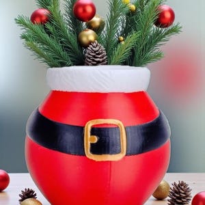 Op de afbeelding: Een feestelijke kerstversiering met een rode kerstman-vaas met een zwarte riem en een gouden gesp. De vaas is gevuld met groene dennentakken, rode en gouden ballen en een dennenappel. Een vakantie-middelpunt.