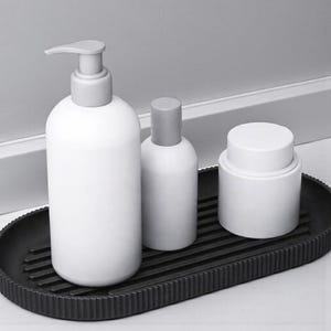 May include: A white soap dispenser, a small bottle, and a jar with gray lids sit on a black, ribbed oval tray. The items are arranged on a white surface, with a white wall and trim in the background.