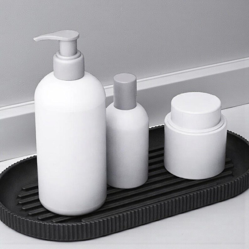 May include: A white soap dispenser, a small bottle, and a jar with gray lids sit on a black, ribbed oval tray. The items are arranged on a white surface, with a white wall and trim in the background.