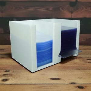 May include: A white acrylic desk organizer with two compartments. One compartment holds a stack of blue circular items, and the other holds a stack of blue rectangular items. The organizer sits on a wooden surface.