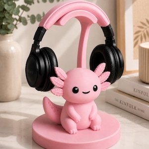 May include: A pink axolotl-shaped headphone stand with a circular base. The stand holds black headphones with the word "SONY" visible. The axolotl has a smiling face and feathery gills. The stand is on a white surface.