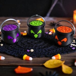 May include: Three black Halloween cauldron decorations with different colored contents: purple, green, and orange. Each cauldron has a silver handle and a jack-o'-lantern face design. Candy corn and plastic spiders are scattered around the cauldrons on a wooden surface.