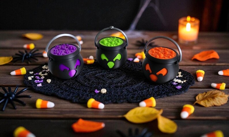 May include: Three black Halloween cauldron decorations with different colored contents: purple, green, and orange. Each cauldron has a silver handle and a jack-o'-lantern face design. Candy corn and plastic spiders are scattered around the cauldrons on a wooden surface.