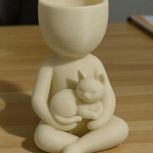 May include: A cream-coloured planter in the form of a seated person holding a sleeping cat. The top of the figure is a large, open bowl for planting. The cat is curled up, adding a touch of serenity to the design.
