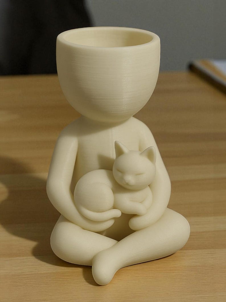 May include: A cream-coloured planter in the form of a seated person holding a sleeping cat. The top of the figure is a large, open bowl for planting. The cat is curled up, adding a touch of serenity to the design.