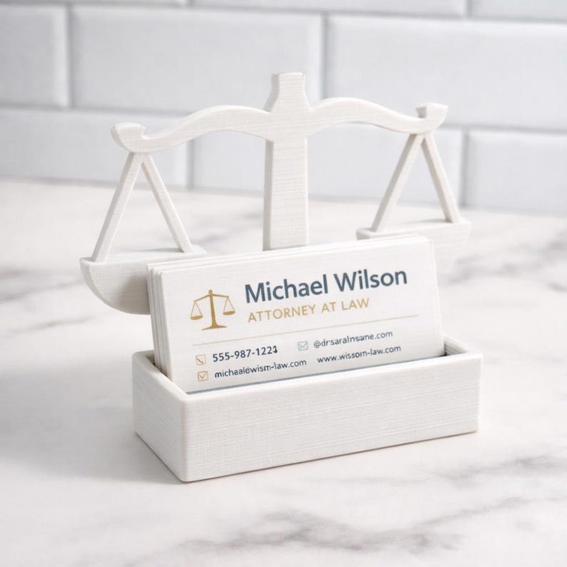 Puede incluir: Un tarjetero de madera blanco con forma de balanza, con tarjetas de visita. Las tarjetas muestran el texto "Michael Wilson, Attorney at Law". El soporte tiene una base rectangular y un dise&ntilde;o de balanza.