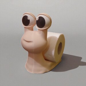 May include: A whimsical, light pink snail-shaped toilet paper holder. The snail has large, cartoonish eyes with brown pupils and a smiling face. A roll of white toilet paper is held in the snail's shell. The holder is on a gray surface.