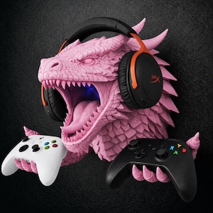 Op de afbeelding: Een roze drakenkop als wandhouder met een textuur van schubben en een open bek. De draak draagt een zwart-oranje koptelefoon en houdt met zijn klauwen een witte gamecontroller aan de linkerkant en een zwarte aan de rechterkant vast.