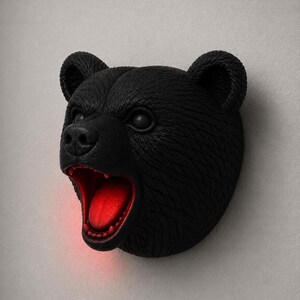 May include: A black bear head wall decor with an open mouth revealing a red interior. The bear has textured fur and is mounted on a light gray wall. The bear's mouth is open, and the interior is illuminated with a red light.