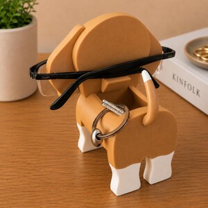 May include: A whimsical dog-shaped desk organizer in tan, white, and brown. The organizer holds eyeglasses and a set of keys. The dog's ears hold the glasses, and the tail and body hold the keys. A decorative item for a desk.