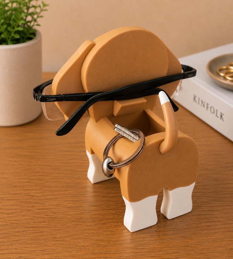 May include: A whimsical dog-shaped desk organizer in tan, white, and brown. The organizer holds eyeglasses and a set of keys. The dog's ears hold the glasses, and the tail and body hold the keys. A decorative item for a desk.